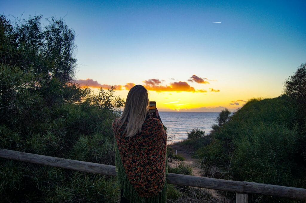 A woman photographs a vibrant sunset over the ocean in Portugal.