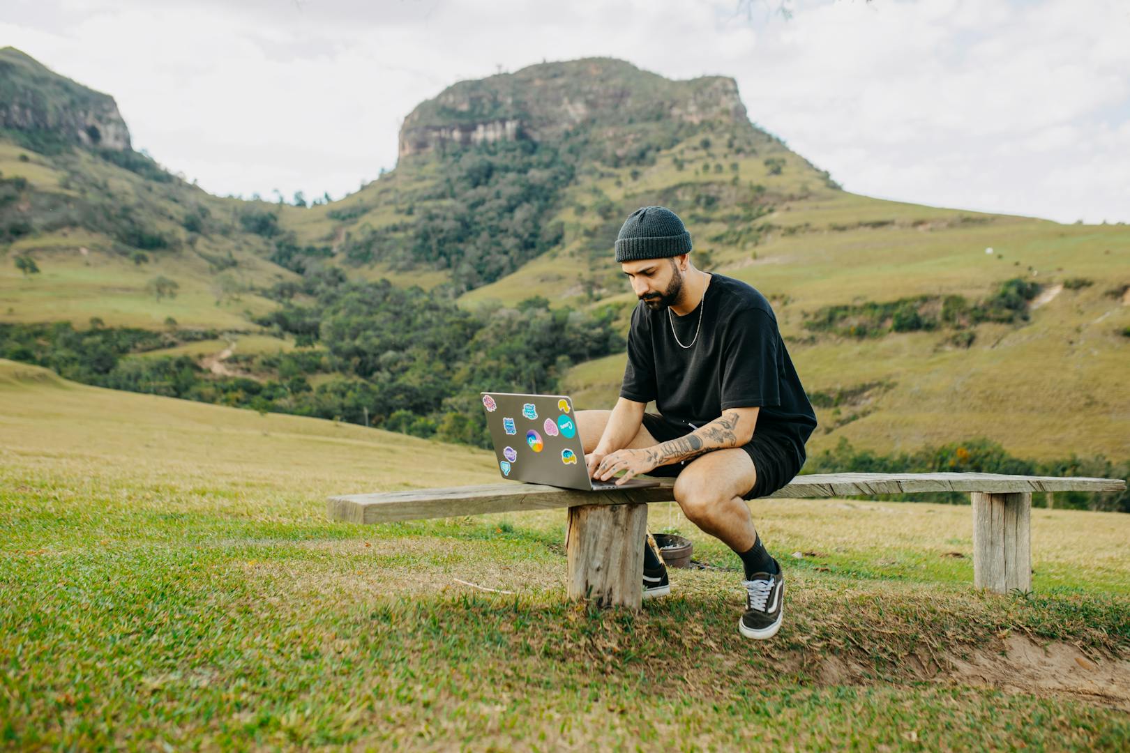 Young man with beard working on laptop outdoors in a scenic mountain setting. Ideal for remote work concepts.