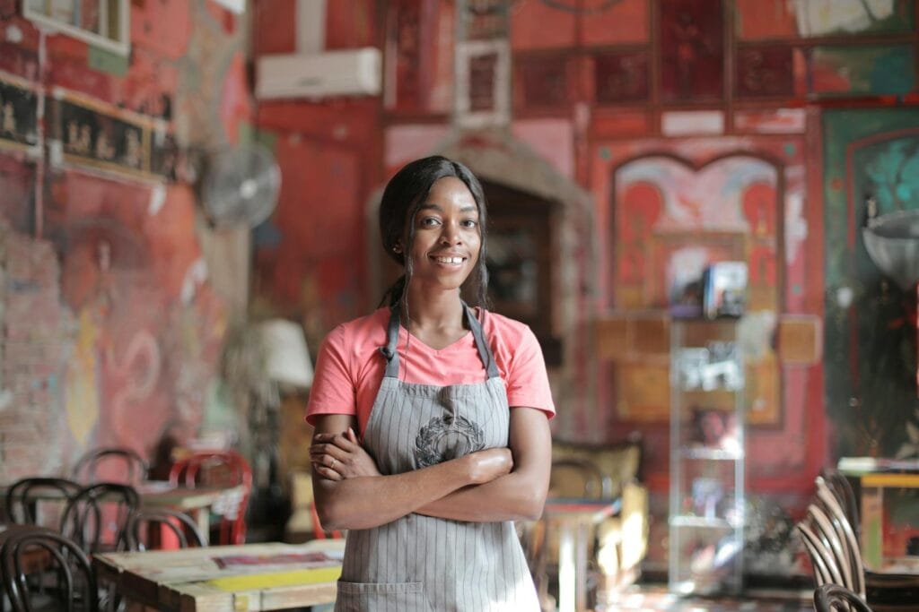 Cheerful waitress in a colorful café with vibrant murals, arms crossed and smiling warmly.