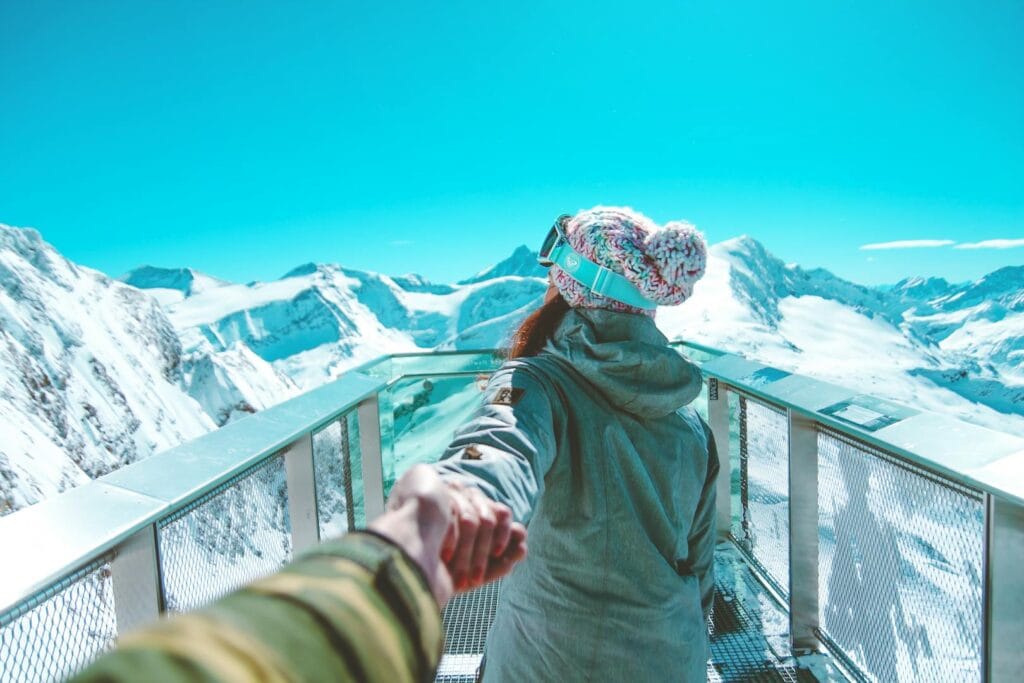 A woman in winter clothing holds a hand and walks on a scenic snow-covered mountain deck in Zell am See.