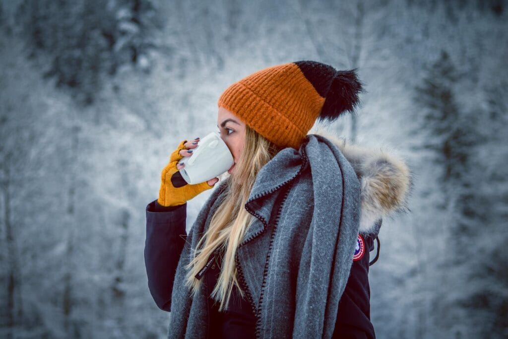 Woman dressed warmly drinking a hot beverage outdoors in a snowy winter setting.