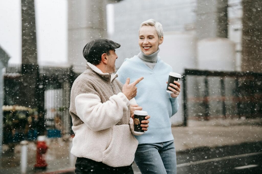 Positive couple crossing road while having conversation and takeaway coffee under snowfall in winter day