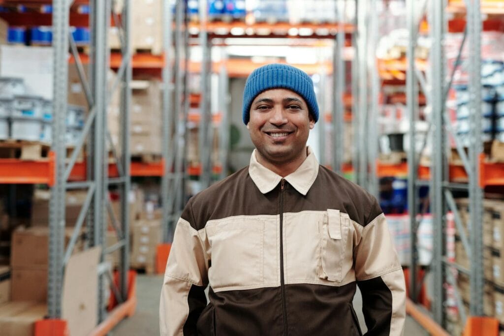 A cheerful warehouse worker wearing a blue beanie in a storage facility.