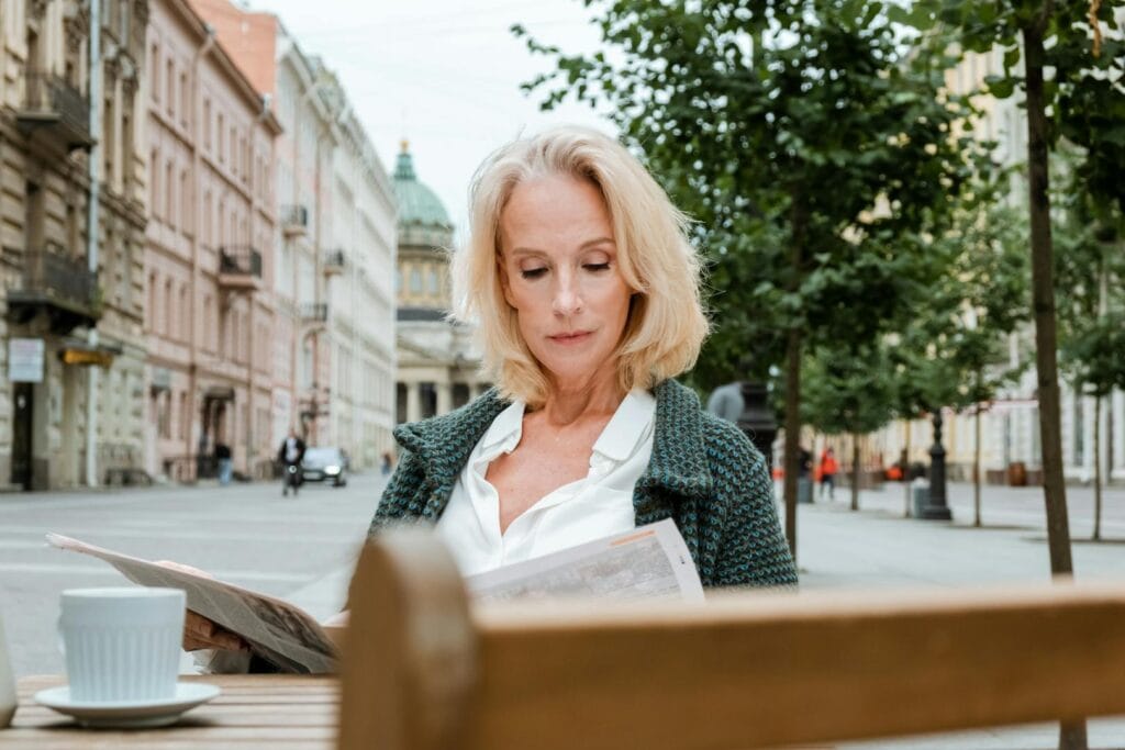 A mature woman reads a newspaper at an outdoor cafe in an urban setting.