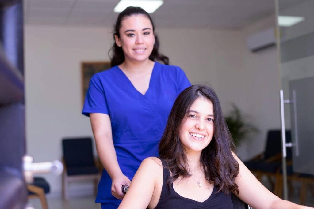 A nurse in blue scrubs assists a smiling patient in a wheelchair indoors.