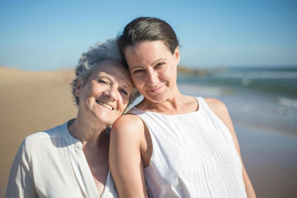 A senior mother and her adult daughter smiling together on a sunny beach in Portugal.