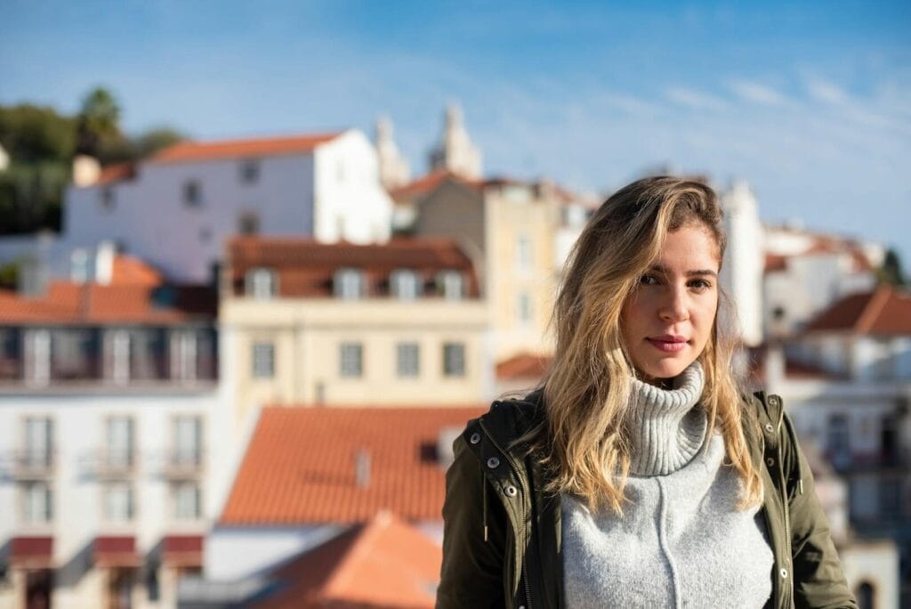 Woman in turtleneck stands outdoors in sunny urban setting. Blurred buildings in background.